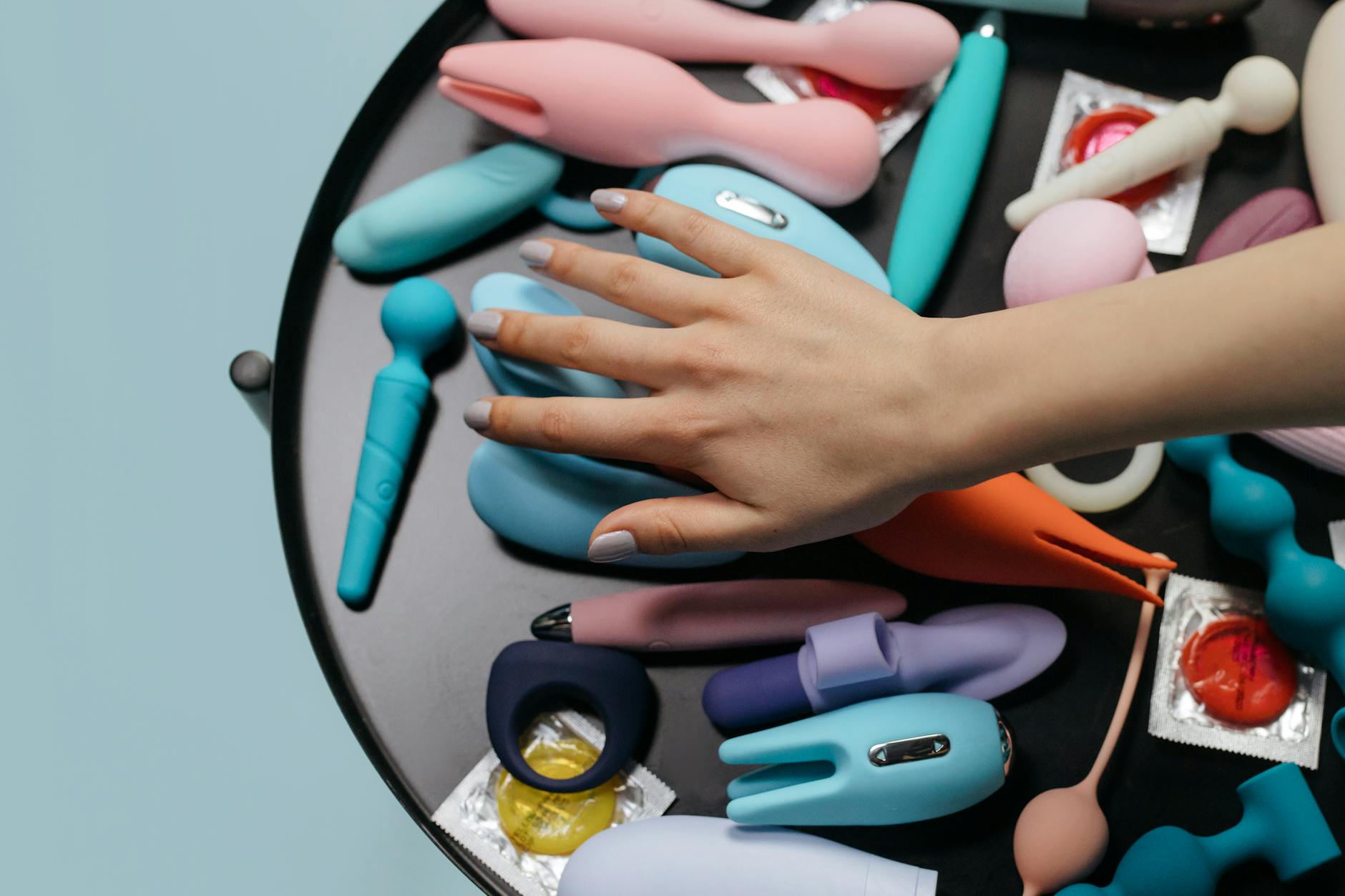 A hand reaching over a variety of colorful sex toys arranged on a table, including clitoral vibrators and suction toys.