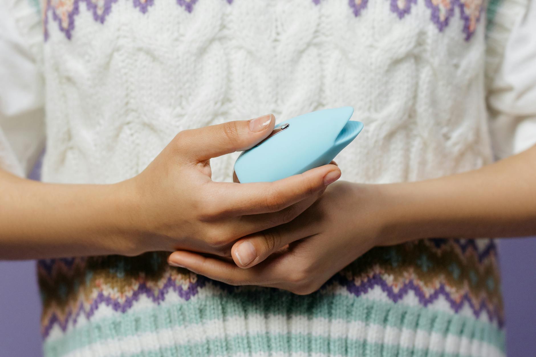 Close-up of hands holding a blue personal massager against a knitted sweater, demonstrating gentle stimulation for sensitive tissue.
